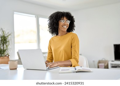 Happy young African American woman wearing eyeglasses using laptop computer technology sitting at home table looking away, hybrid working online, elearning or relaxing in living room. Candid shot. - Powered by Shutterstock