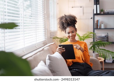 Happy young African American woman sitting on couch at home using digital tablet. - Powered by Shutterstock