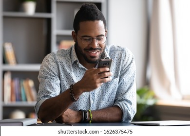 Happy Young African Business Man Student Professional User Hold Smartphone Modern Gadget Texting Message In Social Media App Online Sit At Home Office Desk Using Cell Phone Technology For Surfing Web