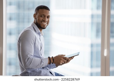 Happy young African business man using online app on tablet for job communication, holding digital gadget, looking at camera, smiling, posing against window city view background. Indoor portrait - Powered by Shutterstock