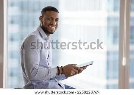 Happy young African business man using online app on tablet for job communication, holding digital gadget, looking at camera, smiling, posing against window city view background. Indoor portrait