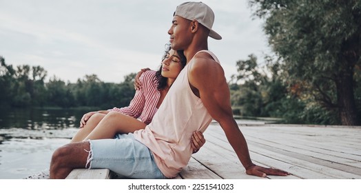 Happy young African couple embracing and smiling while sitting on the pier near the lake - Shutterstock ID 2251541023