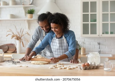 Happy young African couple wearing aprons cooking homemade bakery foods together, kneading, rolling dough for cookies at flour table, talking, laughing. Husband helping wife in home kitchen - Powered by Shutterstock