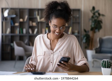 Happy young african ethnic woman in glasses calculating domestic utility bills taxes, managing monthly budget, paying for services online in mobile shopping application, satisfied with enough money. - Shutterstock ID 1978844264