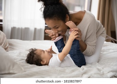 Happy young african ethnicity mum lying on comfortable bed with laughing adorable toddler son daughter, playing tickling indoors. Smiling affectionate biracial mother having fun with kid in bedroom.