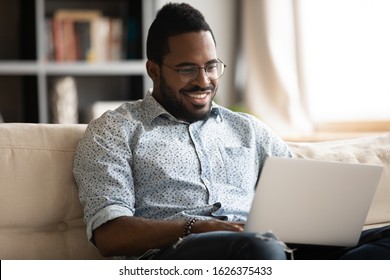 Happy Young African Guy Using Laptop Notebook Relax Sit On Couch With Technology Device, Smiling Afro American Man User Surfing Web On Computer Browsing Internet Work Study Communicate Online At Home