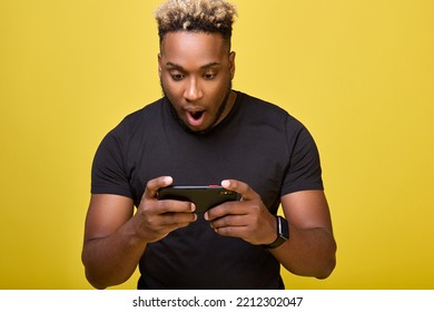 Happy Young African Man With A Fashionable African Haircut, Playing A Video Game On A Smartphone And Having Fun. A Black Guy With His Mouth Open Looks At A Mobile Phone Screen On A Yellow Background