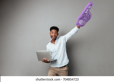 Happy Young African Man Holding Laptop Computer And Foam Finger Isolated Over Gray Background