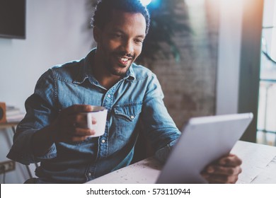 Happy Young African Man Reading World News By Digital Tablet While Sitting At The Table On A Sunny Morning.Concept Of Coworking People Working Home.Blurred Background,flare Effect