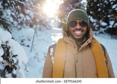 Happy young African man in sunglasses and warm winterwear standing in front of camera against firtrees in snow during chill on winter weekend - Powered by Shutterstock