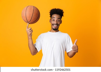 Happy young african man in t-shirt standing isolated over yellow background, playing basketball