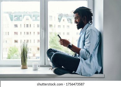 Happy young African man using smart phone and smiling while sitting on the window sill indoors     
