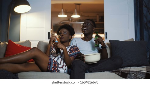 Happy young African positive friends couple watching TV at home on the couch, eating snacks and laughing slow motion.