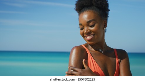 A happy young african woman is applying a sunscreen or sun tanning lotion to take care of her skin during a vacation on a beach.