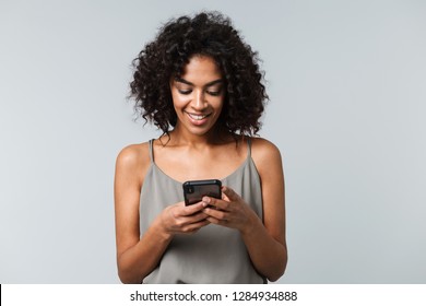 Happy young african woman casually dressed standing isolated over gray background, holding mobile phone