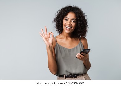 Happy Young African Woman Casually Dressed Standing Isolated Over Gray Background, Holding Mobile Phone, Ok