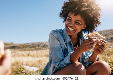 Happy young african woman eating sandwich and smiling. Taking break during country hike.