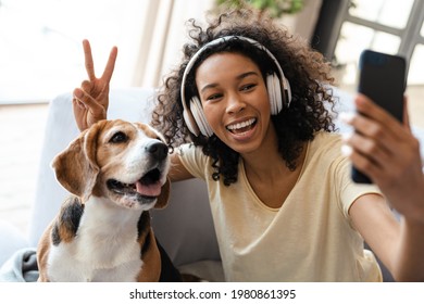 Happy young african woman in headphones relaxing on a couch with her pet dog at home, taing a selfie - Powered by Shutterstock