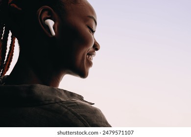 Happy young African woman in her 20s wears wireless earphones, smiling peacefully with her eyes closed. She enjoys listening to music on a white background in a studio, showcasing a relaxed lifestyle - Powered by Shutterstock