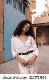 Happy Young African Woman Looking Into Camera With Good Mood, Spends Time Outside In Summer Day. Model Curly Brunette Hair Wears Casual Clothes. Concept Enjoying, Relaxing.