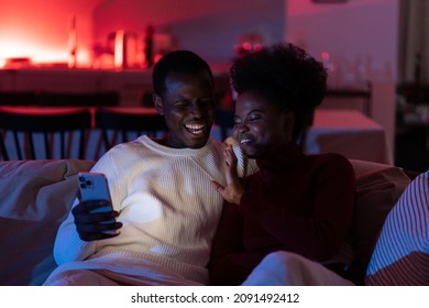 Happy Young African Woman And Man With Smartphone Rest On Couch On Evening Weekend, Smiling Black Couple Look At Phone Screen, Enjoy Video Call With Friends Or Family Via Smartphone. Selective Focus