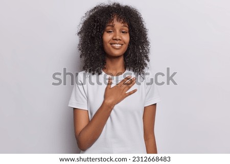 Happy young African woman presses hand to chest demonstrates her appreciation says thank you dressed in casual t shirt smiles pleasantly isolated over white background. Smiling glad female model