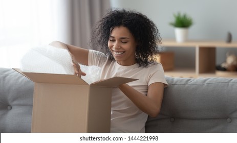 Happy Young African Woman Satisfied Customer Open Parcel Cardboard Box Sit On Sofa At Home, Smiling Black Girl Consumer Unpack Carton Package Receive Good Purchase Postal Shipping Delivery Concept