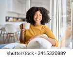 Happy young African woman sitting on comfortable sofa in living room at home. Smiling pretty girl drinking coffee relaxing on couch looking away through window thinking in modern cozy house.