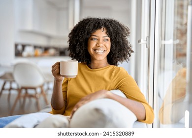 Happy young African woman sitting on comfortable sofa in living room at home. Smiling pretty girl drinking coffee relaxing on couch looking away through window thinking in modern cozy house. - Powered by Shutterstock