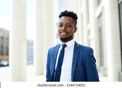 Happy young African-american employee in suit and tie looking at you in urban environment by modern business center