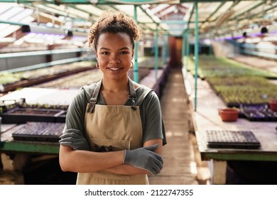 Happy young African-American female garden worker in workwear and protective gloves crossing her arms by chest