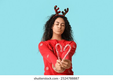 Happy young African-American woman in Christmas sweater with candy canes blowing kiss on blue background - Powered by Shutterstock