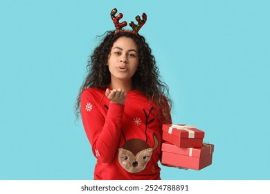 Happy young African-American woman in Christmas sweater with gift boxes blowing kiss on blue background - Powered by Shutterstock