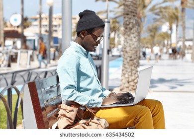Happy young Afro American blogger wearing trendy clothing sitting on bench on urban beach, using laptop pc for while working distantly on new article for online magazine, looking cheerful and inspired