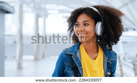 Happy young afro woman listening to playlist music with wireless headphones while wearing yellow shirt and jeans jacket outdoor