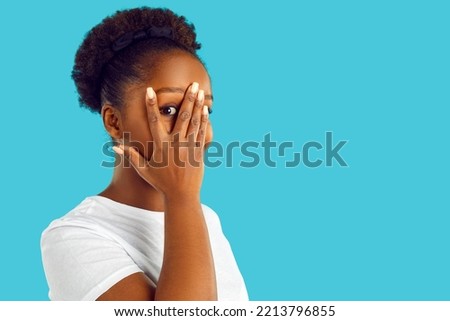 Happy young American woman isolated on blue empty blank advertising copy space background feeling shy, bashful and embarrassed, covering her face with her hand and looking through fingers