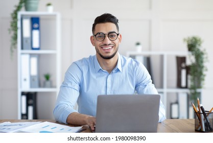 Happy Young Arab Businessman In Glasses Sitting At Desk, Working On Laptop At Company Office. Successful Smiling CEO Having Online Job, Typing On Pc Keyboard, Having Video Call