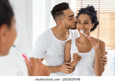 Happy Young Arab Couple Brushing Teeth In Bathroom, Loving Man Kissing His Wife, Romantic Middle Eastern Spouses Making Morning Hygiene Together, Bonding At Home, Selective Focus Ar Mirror Reflection