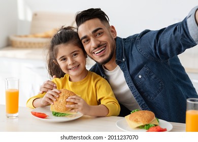 Happy Young Arab Dad Taking Selfie With Little Daughter While Having Lunch Together In Kitchen, Adorable Female Child Enjoying Eating Sandwiches With Father And Smiling At Camera, Closeup