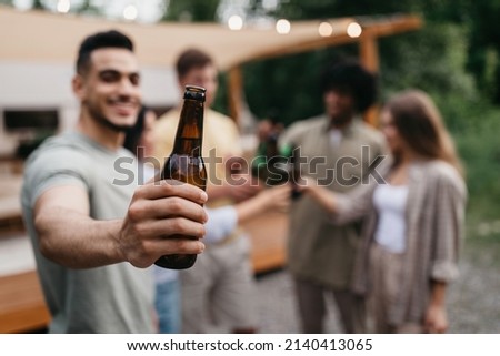 Happy young Arab guy with his diverse friends toasting with beer bottles, drinking alcohol near RV outdoors, selective focus. Millennial people enjoying summer party on camping trip, copy space