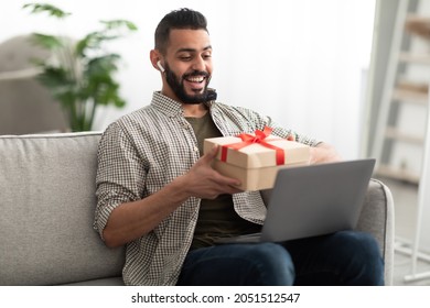 Happy Young Arab Guy Holding Gift Box, Having Video Call Via Laptop, Celebrating Birthday Online With Remote Friends Or Family At Home. Distance Holiday Celebration, Virtual Party Concept