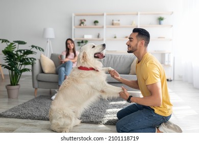 Happy young Arab guy playing with dog, having fun on floor at home, his girlfriend drinking coffee on background, copy space. Interracial married couple with cute pet spending time in living room