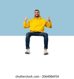 Happy Young Arab Guy Sitting On Huge Horizontal Empty Placard For Advertisement Over Blue Studio Background, Showing Thumb Up And Smiling At Camera, Recommending Something