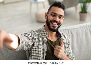 Happy young Arab guy taking selfie and showing thumb up gesture, smiling at mobile phone camera, sitting on sofa at home. Portrait of positive middle Eastern man making photo of himself indoors - Powered by Shutterstock