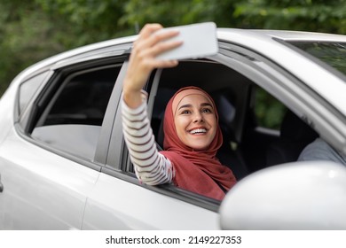 Happy Young Arab Lady In Hijab Takes Selfie On Phone In Open Window In Car, Husband Driving, Outdoor. Travel Blog And Live Stream For Social Networks, Vacation Together And Journey By Auto, Covid-19