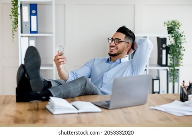 Happy young Arab man in office wear using smartphone, having break, putting feet on table at workplace with laptop pc. Handsome male entrepreneur browsing web, checking messages on phone