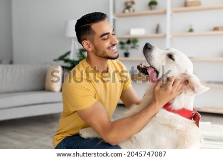Happy young Arab man stroking his adorable dog in living room. Cheerful millennial Eastern guy playing with his pet at home. Human animal friendship concept
