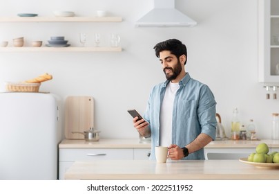 Happy young arab man using smartphone and drinking morning coffee in kitchen, happy millennial guy reading news and enjoying hot drink at home, empty space - Powered by Shutterstock