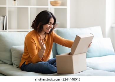 Happy young arab woman opening delivery box at home and looking inside, excited middle eastern lady unboxing parcel with purchases while sitting on couch in cozy living room interior, copy space - Powered by Shutterstock