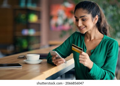 Happy young arabic lady using mobile phone and credit card while having coffee break at cafe, buying something online or paying for goods and services via mobile application, copy space